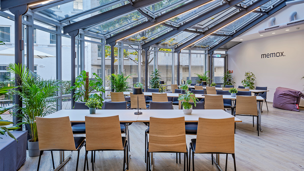 Lichtdurchfluteter Glas-Pavillon mit langen Mittagstischen und Pflanzen