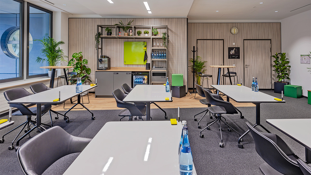 Raum M im Schulungs-Setup mit mehreren Tischreihen, im Hintergrund die Snack- und Getränkeecke